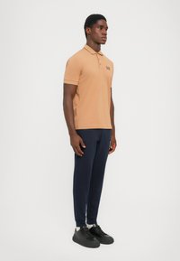 Beige polo-skjorte med korte ermer og marineblå logo, kombinert med marineblå joggebukser og svarte sneakers. Enkel design med klassisk krage.