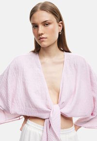 Lys pink cropped top med brede ærmer, foran knytedetalje og struktureret stof. Båret med hvide højtaljede bukser.