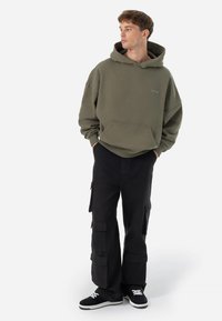 Felpa oversize verde oliva con tasca frontale, abbinata a pantaloni cargo neri con più tasche e sneakers nere con dettagli bianchi.