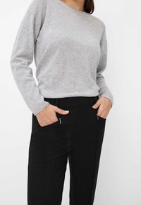 Pull en maille grise avec un col rond, porté avec un pantalon noir doté de deux poches zippées et d'une coupe droite.