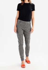 T-shirt noir à manches courtes associé à un pantalon gris texturé et des sandales à talons blocs rouges. Le pantalon présente des détails de coutures et une coupe ajustée.