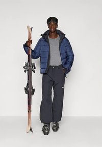 Puchowa granatowa kurtka na szarej koszulce, czarne spodnie narciarskie, trzyma narty z wiązaniami, nosi gogle. Prosty, funkcjonalny strój narciarski.