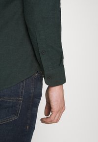 Camisa verde oscuro con tela texturizada, puño abotonado y un dobladillo curvo. Se lleva con vaqueros azul oscuro que presentan costuras contrastantes.