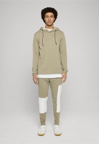 Khaki mikina s kapucí a šnúrkami a bílou vnitřní vrstvou, spárovaná s khaki kalhotami s bílými akcenty. Obojí je vyrobeno z bavlny.