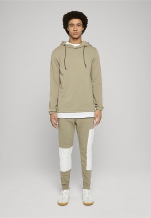 Khakifarbene Kapuzenjacke mit Kordelzügen und einer weißen Innenlage, kombiniert mit khakifarbene Hose mit weißen Akzenten. Beide sind aus Baumwolle gefertigt.