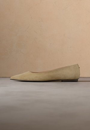 Beige ruskind flad sko med spids tå og lav hæl placeret på en grå overflade mod en beige tekstureret væg.