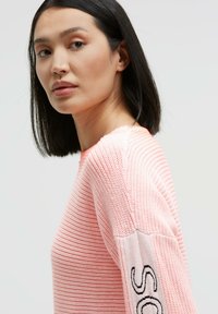 Light pink knitted sweater with textured ribbing, round neckline, and contrasting sleeve featuring embroidered accents.