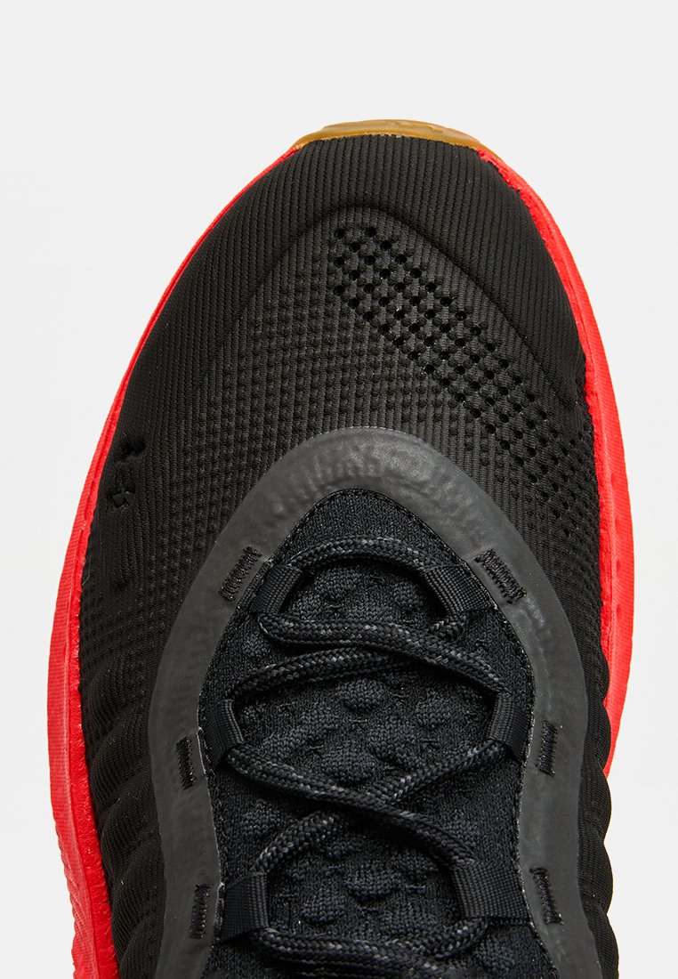 Scarpa sportiva nera con tomaia texture, dettagli rossi sulla suola e lacci neri. Presenta una punta in gomma liscia e sezioni in rete.