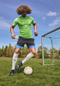 Maglietta sportiva verde neon a maniche corte con un motivo texturizzato, pantaloni corti neri con accenti blu, calze bianche e scarpini da calcio neri su erba.