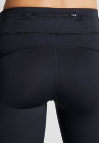 Achteraanzicht van een taille en bovenbenen in zwarte, strakke leggings met een klein, ritszakje op de achterste tailleband.