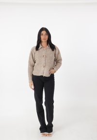 Giacca beige strutturata con colletto, caratterizzata da tre grandi bottoni decorativi, abbinata a pantaloni neri e sandali con punta aperta.