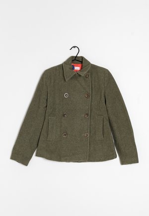 Olivově zelený dvojřadý vlněný kabát se šesti knoflíky, dvěma bočními kapsami a klopovým límcem zavěšený na černém ramínku.