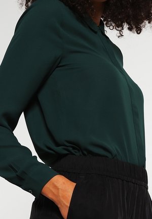 Nainen, joka käyttää tummanvihreää pitkähihainen puseroa, joka on sullottu mustiin joustavaan vyötäröiseen housuihin, yhdellä kädellä taskussa.