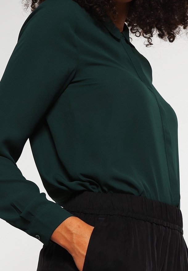 Femme portant un chemisier vert foncé à manches longues, rentré dans un pantalon noir avec une taille élastique, une main dans la poche.