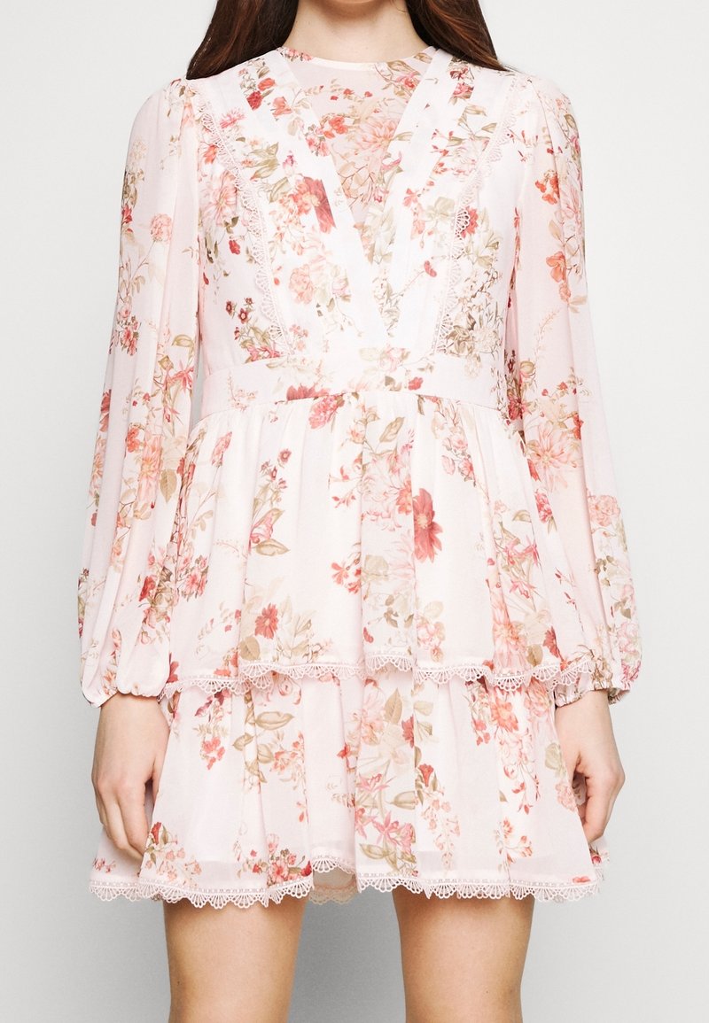 Robe blanche à fleurs avec manches longues bouffantes, jupe à volants, bordure en dentelle et décolleté en V portée par une personne.
