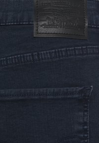 Jean en denim bleu foncé avec une étiquette en cuir comportant un logo en relief. Détails de couture visibles et une poche arrière. Texture lisse.