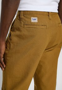 Zbliżenie brązowych spodni roboczych Lee noszonych przez osobę o ciemnej skórze w białej koszuli, z widoczną tylną kieszenią i metką marki.