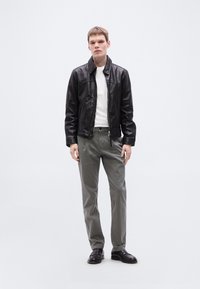 Μαύρο δερμάτινο μπουφάν πάνω από ένα λευκό πουκάμισο, συνδυασμένο με γκρι παντελόνι και μαύρα παπούτσια. Το μοντέλο στέκεται όρθιο μπροστά σε απλό φόντο.