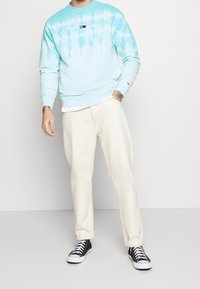 Jasnoniebieska bluza tie-dye z białym logo, zestawiona z kremowymi spodniami i czarnymi trampkami. Swobodny krój, ściągacze na mankietach i dole.