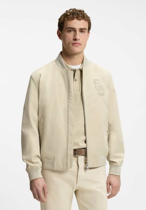 Junger Mann, der eine beige Collegejacke über einem beigen Poloshirt und einer Hose trägt, vor einem schlichten weißen Hintergrund stehend.