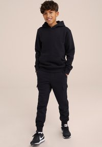 Svart bomullshoodie med ficka framtill, matchad med svarta cargojoggers som har sidofickor. Bärs med svarta sneakers.