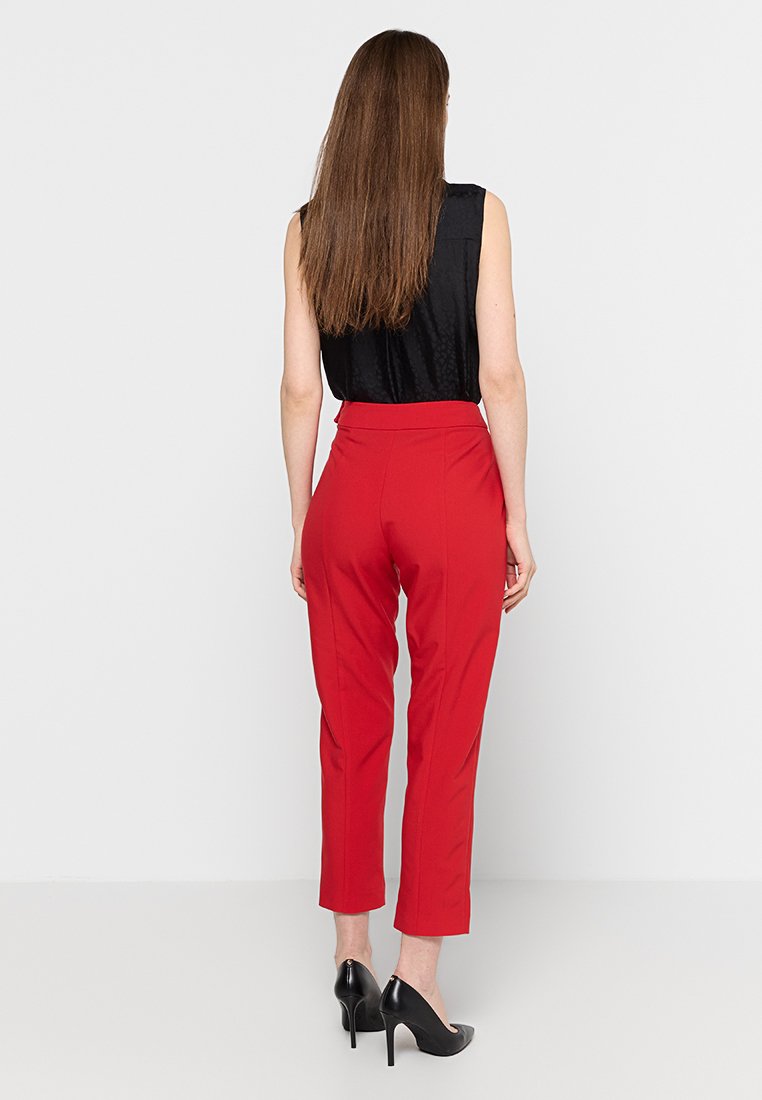 Pantaloni tailored rossi con vita alta, gambe dritte e una texture liscia. Indossati con un top senza maniche nero e tacchi neri.