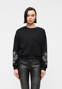Femme portant un sweat-shirt noir avec une broderie florale blanche sur les manches et un pantalon noir texturé, debout devant un fond clair uni.
