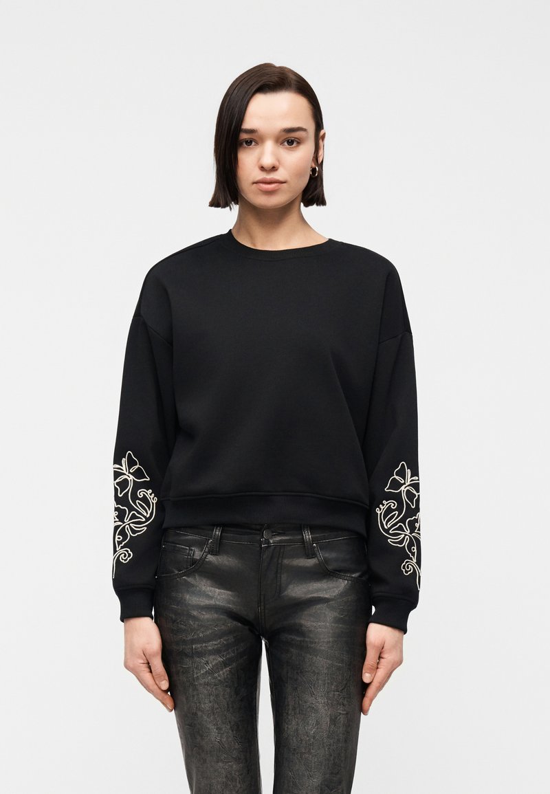 Femme portant un sweat-shirt noir avec une broderie florale blanche sur les manches et un pantalon noir texturé, debout devant un fond clair uni.