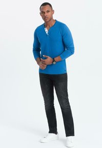 Camicia azzurra a maniche lunghe con dettagli a bottoni, abbinata a jeans scuri e sneakers bianche. Il tessuto sembra morbido e aderente.
