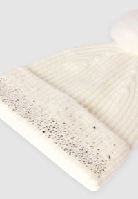 Biała dzianinowa zimowa czapka z pomponem, ozdobiona małymi srebrnymi ćwiekami wzdłuż zakrzywionego brzegu.