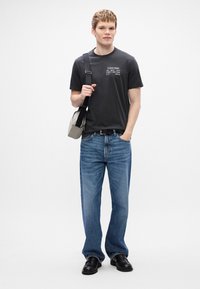 Γκρι βαμβακερό μπλουζάκι με εκτυπωμένο λογότυπο, μπλε τζιν παντελόνι και μαύρα δερμάτινα παπούτσια. Κρατώντας μια μικρή γκρι τσάντα ώμου με λουράκι.