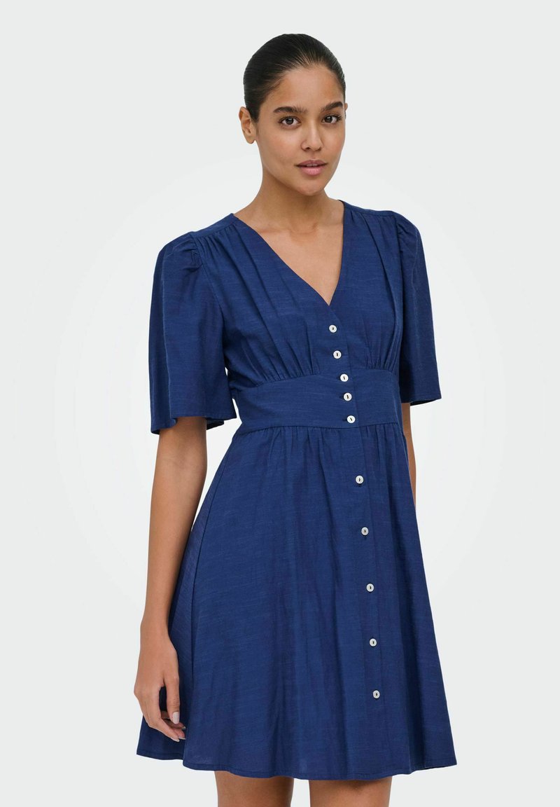 Femme portant une robe bleu marine à manches courtes, col en V, avec des boutons blancs sur le devant, sur un fond uni.