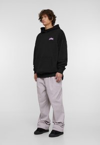 Melns kapučjaka ar rozā logo, brīva piegriezuma, priekšējā kabata, apvienots ar gaiši pelēkiem platajiem biksēm un melniem sporta apaviem, stāv uz vienkārša fona.