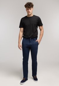 Czarny bawełniany t-shirt, granatowe dżinsy o dopasowanym kroju oraz granatowe sneakersy z brązowymi sznurowadłami. Model stoi na tle o neutralnym kolorze.