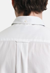 White shirt, cotton material, classic collar, button closure at neck, back pleat detail, smooth texture, no patterns or accents.