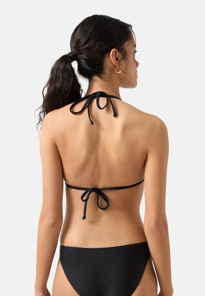 Femme aux cheveux foncés attachés en queue de cheval, portant un haut de bikini noir avec un lien dans le dos et un bas de bikini noir, de dos sur un fond uni.