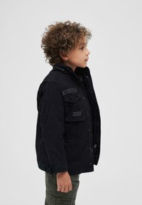 Juodas švarkas su aukštu apykakle, priekiniais užtrauktukais ir keliais kišenėmis. Puošiamas pleistrais ir tekstūruota medžiaga, vilkimo vaiko.