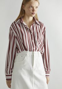Camisa a rayas en rojo oscuro y blanco, de mangas largas, con botones en la parte frontal y un bolsillo en el pecho. Combinada con una falda de mezclilla blanca de cintura alta.