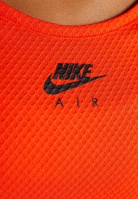 Orange sports top med et tekstureret mønster, der har et sort Nike-logo og "AIR" tekst nedenfor. Runde halsudskæring og korte ærmer.