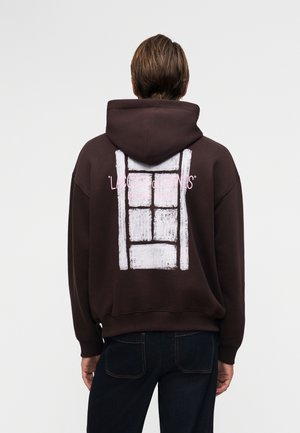 Persoon van achteren gezien, draagt een donkerbruine hoodie met een wit rechthoekig grafisch ontwerp en roze tekst op de achterkant, gecombineerd met een donkere spijkerbroek.