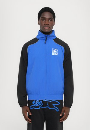 Blauwe en zwarte zip-up jas met hoge kraag, voorzien van een logo patch en contrasterende stiksels. Gladde textuur en sportief ontwerp.