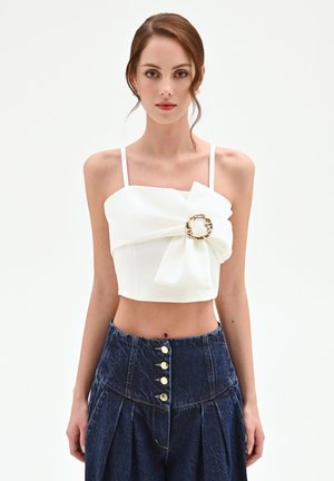 Donna che indossa un top corto bianco con un grande fiocco e una fibbia rotonda dorata, abbinato a pantaloni in denim scuro a vita alta con bottoni dorati.