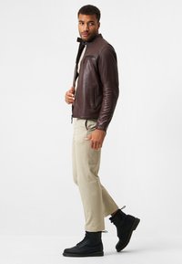 Brun läderjacka med slim fit, dragkedja på bröstfickan, ribbade ärmslut, tillsammans med beige byxor och svarta snörstövlar.