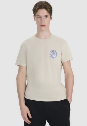 Młody mężczyzna o jasnobrązowych włosach, ubrany w beżowy t-shirt z niebieskim okrągłym logo „Everyday Peace” i czarne spodnie, stoi na jednolitym tle.