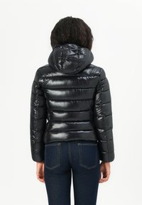 Zwarte pufferjack met een glanzende afwerking, capuchon en horizontale quilt-details. Gecombineerd met donkerblauwe jeans en een relaxte pasvorm.