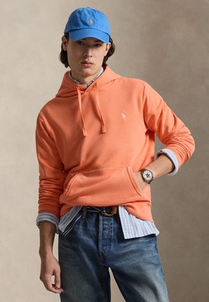 Mlad moški, ki nosi modro kapo, oranžen pulover s kapuco čez modro črtasto srajco, kavbojke in ročno uro, stoji pred nevtralnim ozadjem.