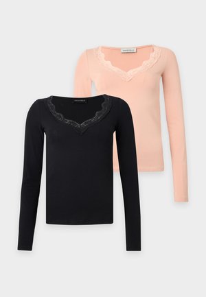 Femme portant un haut noir à manches longues avec un col en dentelle et un pantalon à jambes larges, tenant un sac à main noir, debout sur un fond blanc uni.