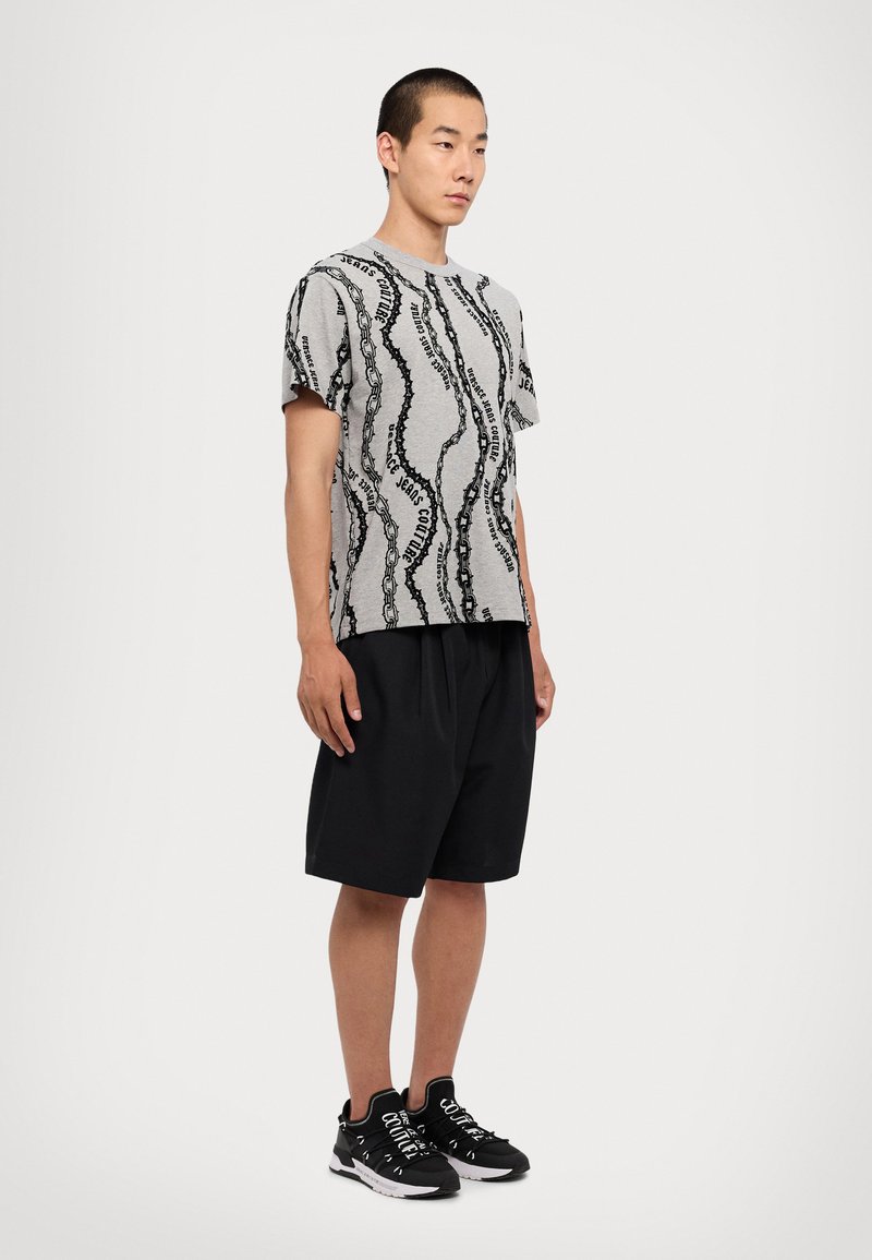 Graues kurzärmliges Shirt mit schwarzem wellenförmigem Textmuster, kombiniert mit schwarzen Shorts und schwarzen Sportschuhen mit weißen Akzenten.