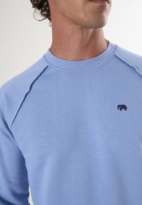Felpa azzurra chiara realizzata in tessuto morbido, con collo rotondo e maniche raglan; include un piccolo logo di un elefante nero sul petto.