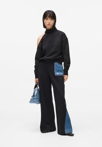 Sudadera negra con escote caído y cuello alto, combinada con pantalones de pierna ancha que tienen un parche de denim. Los accesorios incluyen un pequeño bolso azul.
